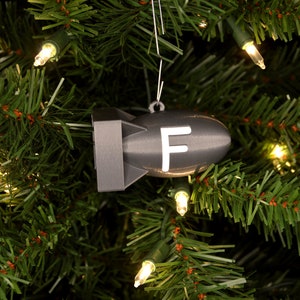 May include: A gray Christmas ornament shaped like a bomb with a white "F" on the side, hanging on a green Christmas tree with white lights.