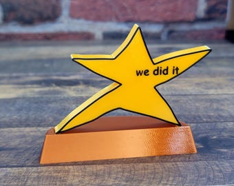 May include: A yellow star-shaped trophy with a black outline and the text "we did it" on the front. The trophy is sitting on a brown base.