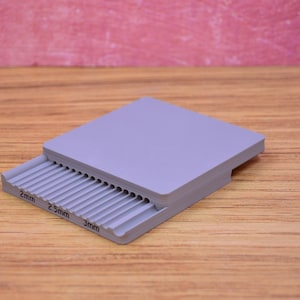 May include: A light purple plastic gauge tool featuring a series of parallel grooves with printed labels for 2mm, 2.5mm, and 3mm measurements. The rectangular device has a flat top surface and a stepped edge where the grooved section is located.