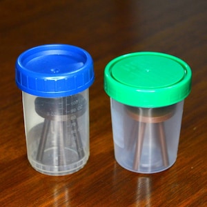 May include: Two clear plastic containers with lids. The container on the left has a blue lid and the container on the right has a green lid. Both containers have a small metal tripod inside.
