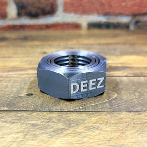 May include: A silver metal nut with the word "DEEZ" engraved on the side. The nut is made of multiple layers and is sitting on a wooden surface.