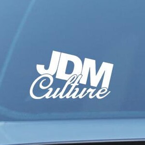 Puede incluir: Adhesivo de vinilo blanco con el texto "JDM Culture" pegado en la ventana de un coche. El diseño presenta "JDM" en una fuente audaz y en bloque, con "Culture" en escritura cursiva debajo.