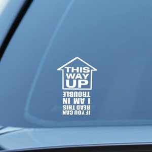 May include: White vinyl sticker with the text "This Way Up" and the words "If you can read this I am in trouble" on a blue car window.