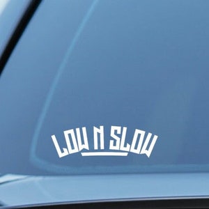 Puede incluir: Calcomanía de vinilo blanco con el texto "LOW N SLOW" en una fuente estilizada, aplicada a la ventana trasera de un vehículo. La calcomanía está centrada y tiene una estética limpia y moderna. La ventana del coche está tintada.
