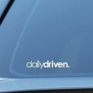 Könnte beinhalten: Eine Heckscheibe eines Autos mit einem weißen Vinyl-Aufkleber, auf dem "dailydriven." steht.