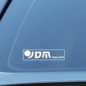 Może przedstawiać: Biała naklejka na szybę samochodu z kółkiem i napisem "JDM squad" w kolorze białym.