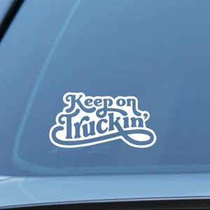 Può includere: Adesivo in vinile bianco con il testo "Keep on Truckin'" in un font retrò. L'adesivo è applicato sul lunotto posteriore di un'auto blu.