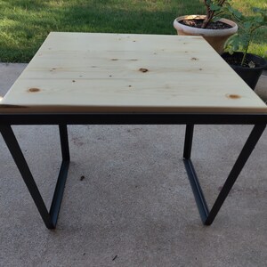 May include: A square wooden table top with a black metal frame. The table top is light wood with a natural finish. The frame is made of simple, straight lines.