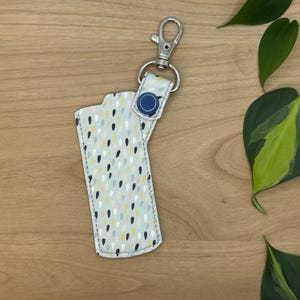May include: A white keychain with a blue button and a silver clip. The keychain has a pattern of small, colorful dots and teardrop shapes.