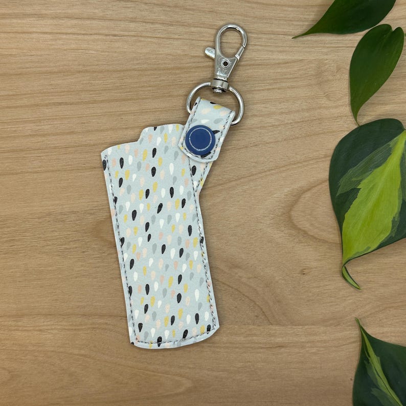 May include: A white keychain with a blue button and a silver clip. The keychain has a pattern of small, colorful dots and teardrop shapes.