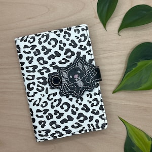 May include: A black and white leopard print wallet with a black bat applique. The bat has pink stitching for the eyes and nose.