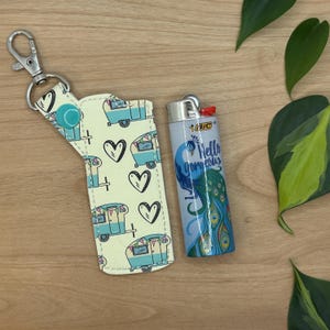 May include: A yellow and blue patterned key chain with a small, blue and white camper van design. The key chain has a silver metal ring and a blue button. The key chain is next to a blue lighter with a peacock design and the text "Hello Gorgeous".