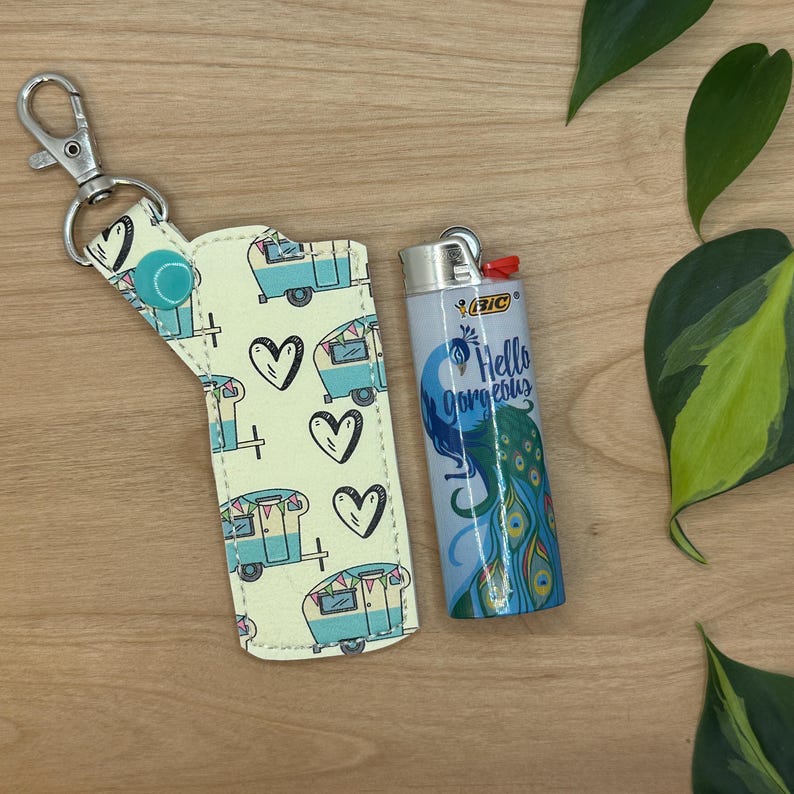 May include: A yellow and blue patterned key chain with a small, blue and white camper van design. The key chain has a silver metal ring and a blue button. The key chain is next to a blue lighter with a peacock design and the text "Hello Gorgeous".