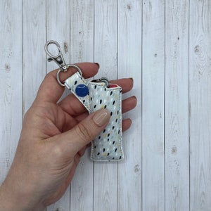 May include: A white fabric lighter case with a colorful abstract pattern. The case has a silver keychain attached and a blue button.