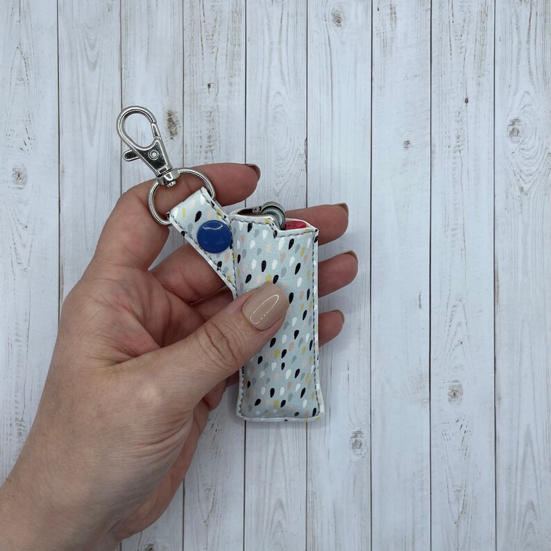 May include: A white fabric lighter case with a colorful abstract pattern. The case has a silver keychain attached and a blue button.
