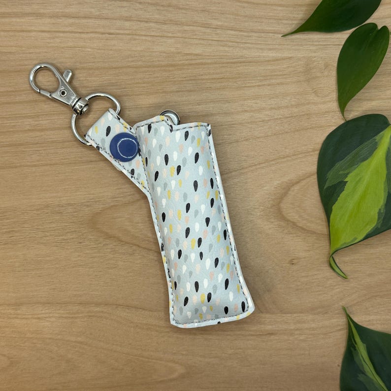 May include: A white zippered pouch with a grey, black, and yellow raindrop pattern. The pouch has a blue button and a silver key ring.