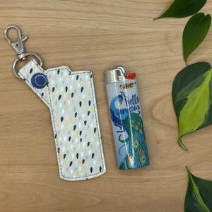 May include: A white and gray patterned lighter case with a silver key chain. The case is next to a silver lighter with a blue and green peacock design and the text "Hello gorgeous".