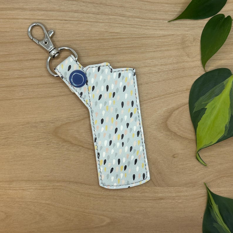 May include: A gray fabric key chain with a blue button and a white, black, and yellow raindrop pattern. The key chain is attached to a silver metal key ring.