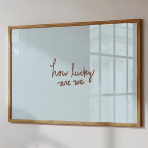 May include: A framed wall art piece with the phrase "how lucky are we" in a handwritten style. The text is in a warm brown color against a light blue background. The frame is made of light brown wood.