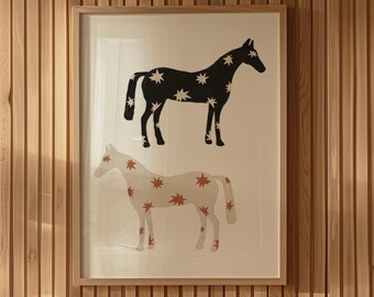 May include: A framed print of two horses with a star pattern. The top horse is black with white stars, and the bottom horse is white with orange stars. The print is hanging on a wood panel wall. A beige ceramic lamp sits on a table with two books.