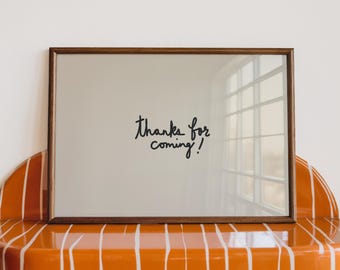 May include: A framed mirror with the text "Thanks for coming!" sits on an orange shelf with white vertical stripes. The shelf has a rounded edge and is mounted on a white wall. The mirror has a dark brown frame.