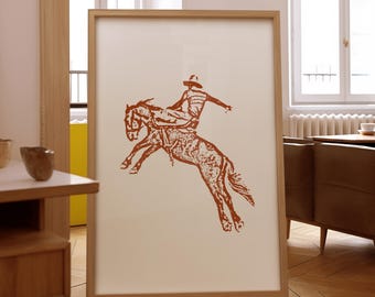 May include: A framed print of a red-brown illustration of a cowboy riding a bucking horse. The print is on a white background and is framed in a light wood frame.