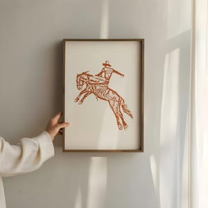 May include: A framed print of a brown silhouette of a cowboy riding a bucking horse. The print is on a white background.