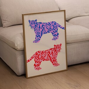 Op de afbeelding: Een ingelijst drukwerk met twee gestileerde jaguars. De bovenste jaguar is roze met blauwe vlekken en de onderste jaguar is rood met roze vlekken. Beide jaguars kijken naar voren met hun mond dicht.