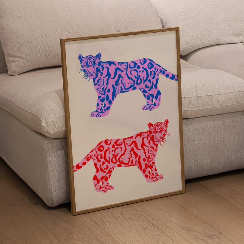 Op de afbeelding: Een ingelijst drukwerk met twee gestileerde jaguars. De bovenste jaguar is roze met blauwe vlekken en de onderste jaguar is rood met roze vlekken. Beide jaguars kijken naar voren met hun mond dicht.