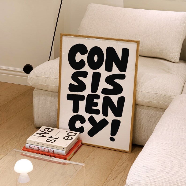 May include: A framed print with the word "CONSISTENCY" in large, black, bold letters on a white background. The print is leaning against a white couch.