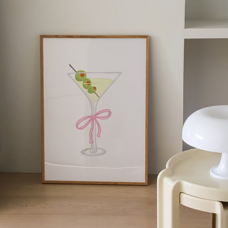 May include: A framed print depicting a martini glass. The glass contains a yellow liquid and is decorated with three green olives on a cocktail stick. A pink bow is tied around the base of the glass.