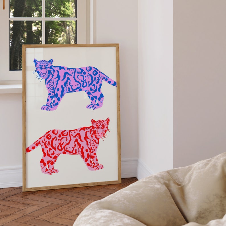Op de afbeelding: Een ingelijste print met twee gestileerde illustraties van roze en blauwe jaguars met zwarte contouren. De jaguars kijken naar voren met hun mond dicht. De achtergrond is wit.
