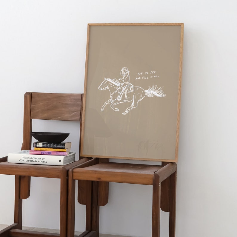 Pu&ograve; includere: Una stampa incorniciata con uno sfondo beige e un disegno a linea bianca di una persona a cavallo. Il testo "Off to see and feel it all" &egrave; scritto in bianco sotto l'immagine.