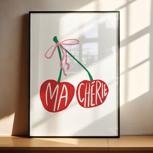 May include: A framed print with a white background featuring two red cherries with green stems and a pink ribbon. The cherries have the words "MA CHERIE" written on them.