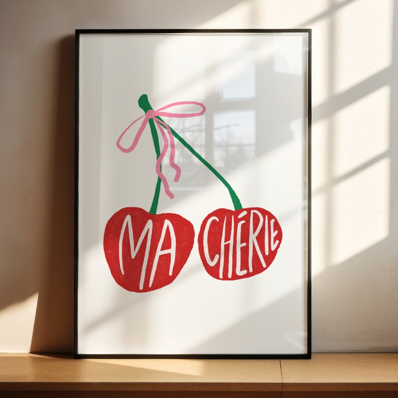 May include: A framed print with a white background featuring two red cherries with green stems and a pink ribbon. The cherries have the words "MA CHERIE" written on them.