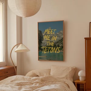 May include: A framed print with a mountain scene and the text "Meet me in the Tetons" in yellow lettering. The print is hanging on a wall in a bedroom with a bed and a lamp.