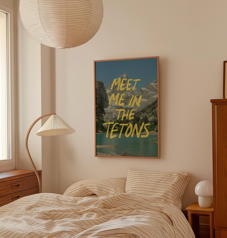 May include: A framed print with a mountain scene and the text "Meet me in the Tetons" in yellow lettering. The print is hanging on a wall in a bedroom with a bed and a lamp.
