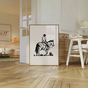 Op de afbeelding: Zwart-witte lijntekening van een cowboy die op een paard rijdt. De print is ingelijst en hangt aan een witte muur.