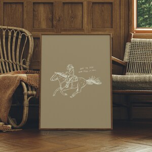 Pu&ograve; includere: Una stampa incorniciata beige con un disegno a linea di una persona a cavallo. Il testo "OFF TO SEE AND FEEL IT ALL" &egrave; scritto in bianco sulla stampa.