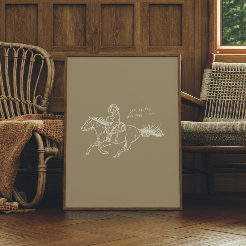 Pu&ograve; includere: Una stampa incorniciata beige con un disegno a linea di una persona a cavallo. Il testo "OFF TO SEE AND FEEL IT ALL" &egrave; scritto in bianco sulla stampa.