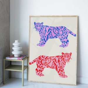 Op de afbeelding: Een ingelijste print met twee gestileerde illustraties van roze en blauwe jaguars met zwarte contouren. De jaguars zijn afgebeeld in een speelse, grafische stijl.