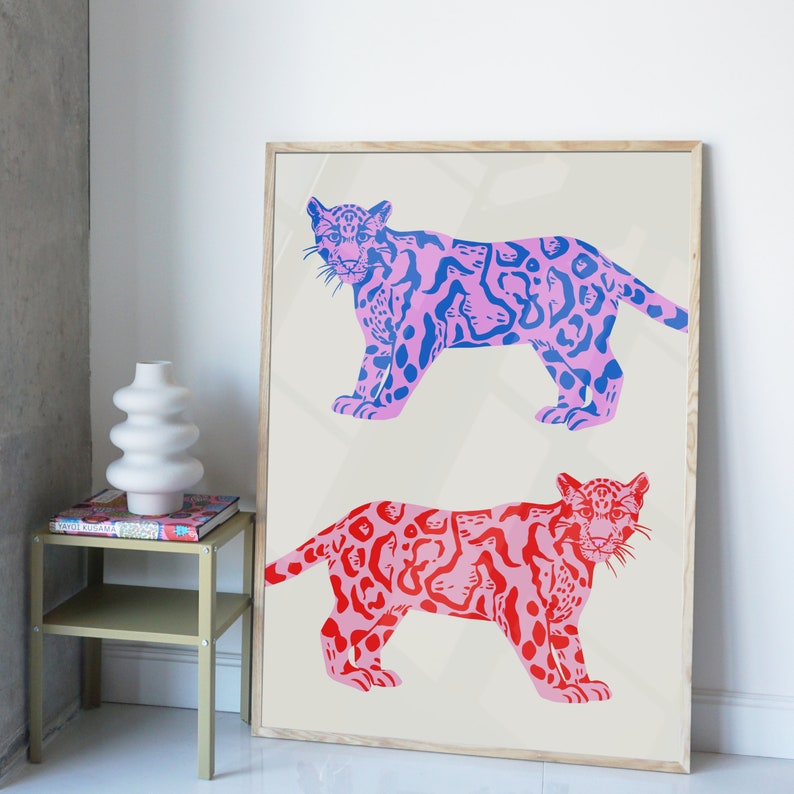 Op de afbeelding: Een ingelijste print met twee gestileerde illustraties van roze en blauwe jaguars met zwarte contouren. De jaguars zijn afgebeeld in een speelse, grafische stijl.