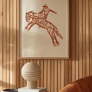 May include: A framed print of a cowboy riding a bucking horse. The print is in shades of brown and white and is set against a wood panel wall.