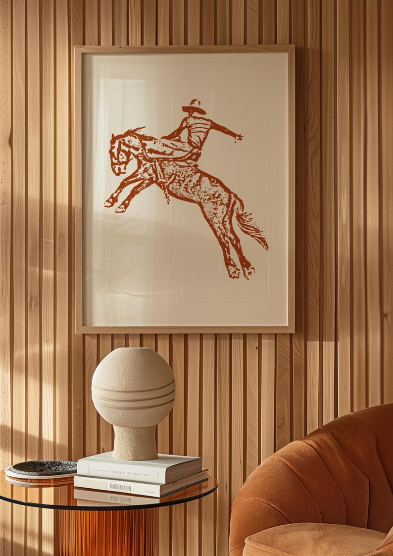 May include: A framed print of a cowboy riding a bucking horse. The print is in shades of brown and white and is set against a wood panel wall.