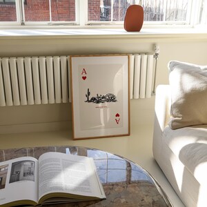 Op de afbeelding: Een ingelijst afdruk van de Aas van harten speelkaart met een woestijnlandschap illustratie. De kaart is ingelijst in een licht houten lijst en hangt aan een witte muur. De muur bevindt zich naast een witte radiator en een witte bank.