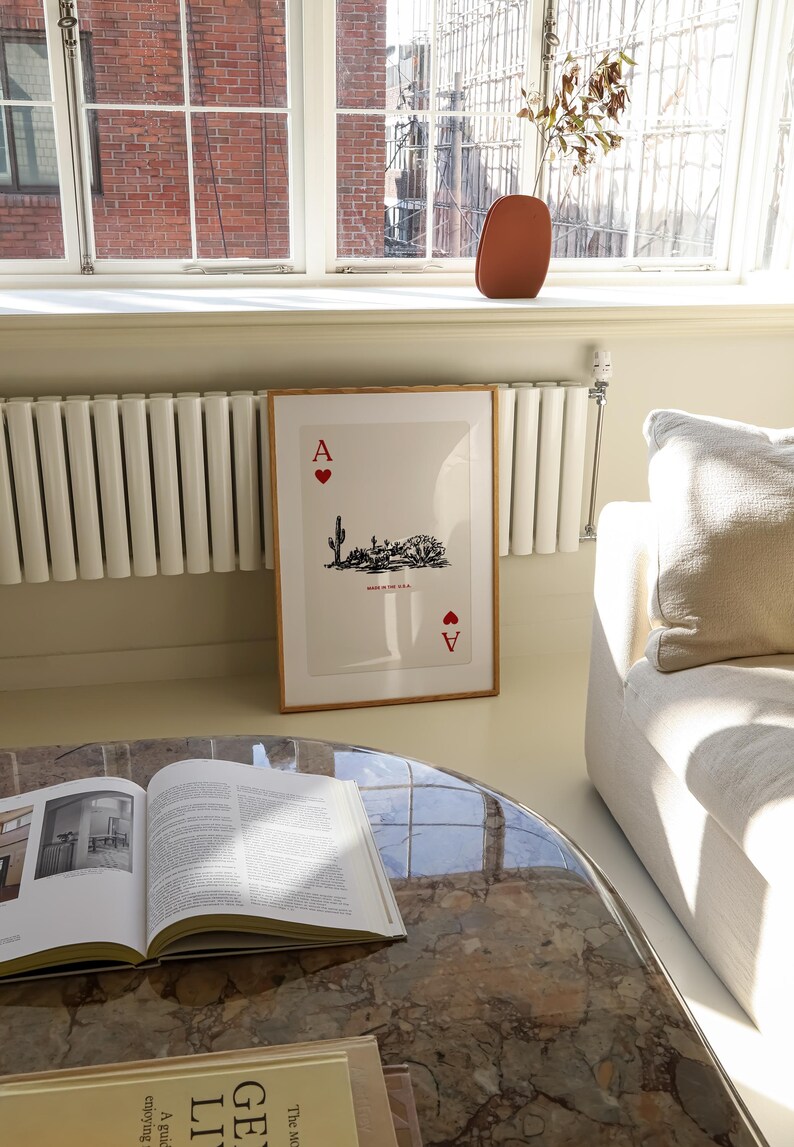 Op de afbeelding: Een ingelijst afdruk van de Aas van harten speelkaart met een woestijnlandschap illustratie. De kaart is ingelijst in een licht houten lijst en hangt aan een witte muur. De muur bevindt zich naast een witte radiator en een witte bank.