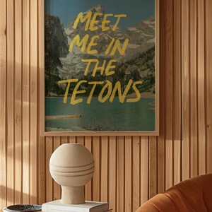 May include: A framed print with a mountain landscape and the text "Meet me in the Tetons" in yellow paint. The print is hanging on a wood panel wall. A beige ceramic lamp sits on a glass table with books.