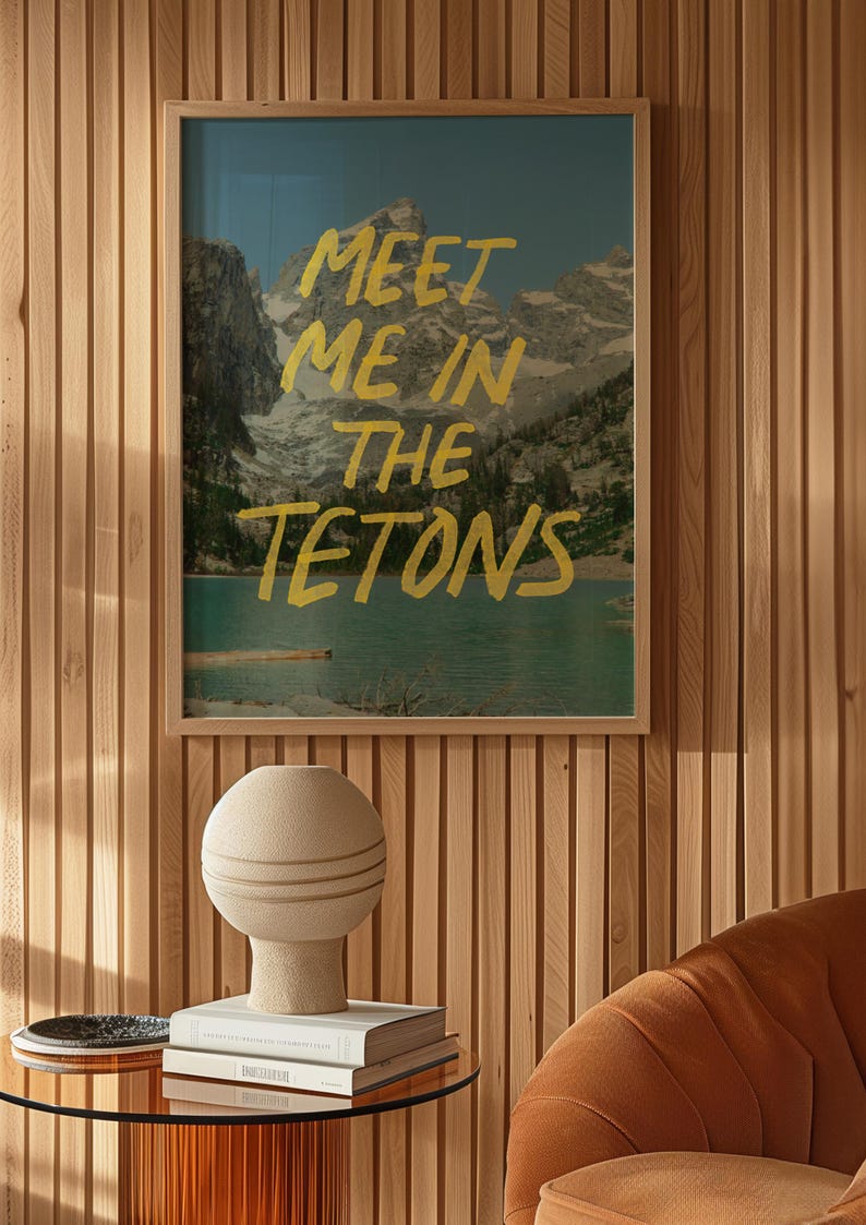 May include: A framed print with a mountain landscape and the text "Meet me in the Tetons" in yellow paint. The print is hanging on a wood panel wall. A beige ceramic lamp sits on a glass table with books.