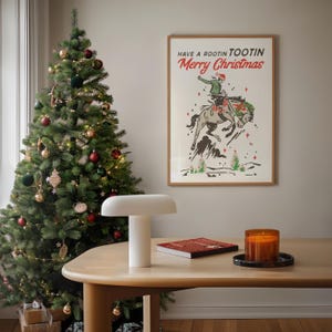 Puede incluir: Una escena navide&ntilde;a festiva con un &aacute;rbol decorado con adornos y luces. Un cuadro enmarcado con un tema de vaquero que dice "Have a Rootin' Tootin' Merry Christmas" cuelga de la pared. Una l&aacute;mpara blanca, un libro y una vela est&aacute;n sobre una mesa de madera.