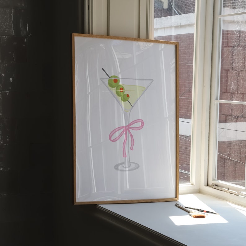 May include: A framed print depicting a martini glass with three olives on a cocktail stick. The glass is adorned with a pink bow. The artwork is displayed near a window.